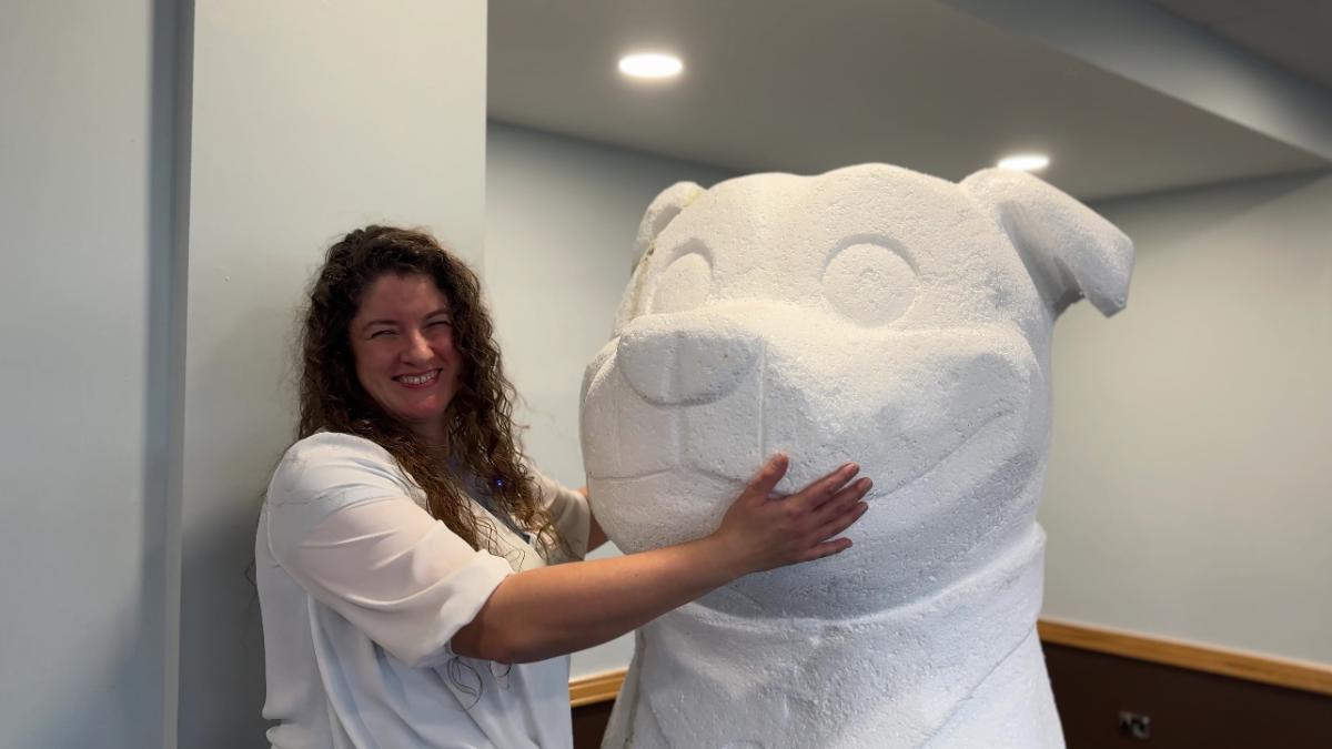 A woman with curly brown hair smiling as she hugs a huge white sculpture of a Staffordshire Bull Terrier dog